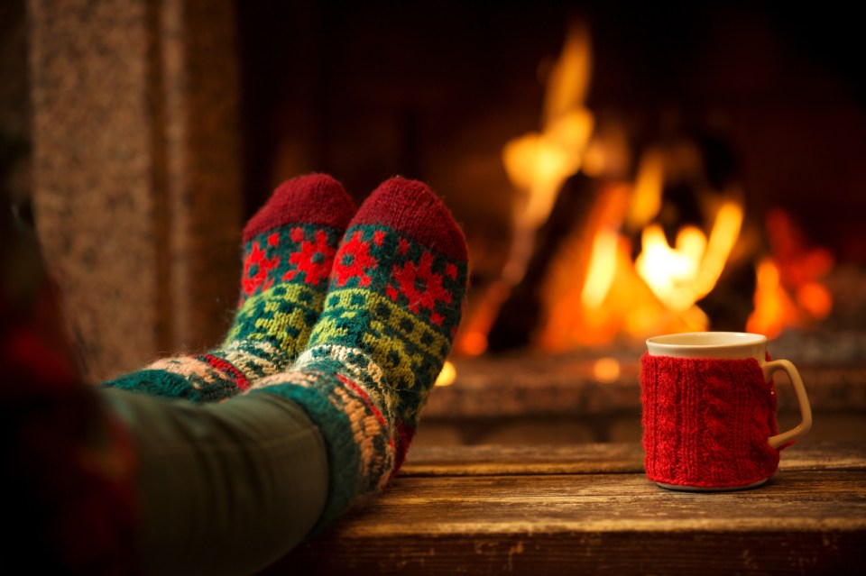 Feet in woollen socks by the Christmas fireplace. Woman relaxes by warm fire with a cup of hot drink and warming up her feet in woollen socks. Close up on feet. Winter and Christmas holidays concept.
